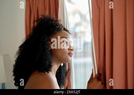 Eine Frau, die aus einem Fenster mit Vorhängen blickt und einen nachdenklichen Ausdruck zeigt. Stockfoto