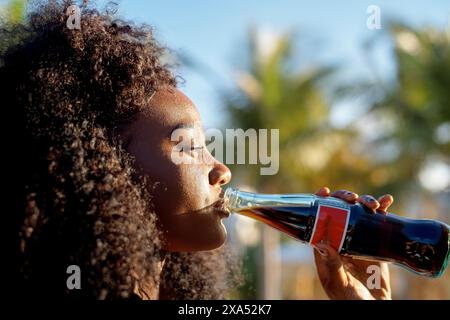 Seitenansicht einer jungen Frau, die draußen einen Drink aus einer Glasflasche genießt, wobei das Sonnenlicht ihr Profil unterstreicht. Stockfoto