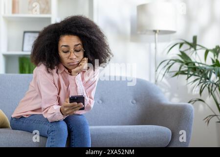 Junge Frau mit Brille, die sich langweilig fühlt und auf das Smartphone schaut, während sie auf einem grauen Sofa in gemütlicher Atmosphäre sitzt. Stockfoto