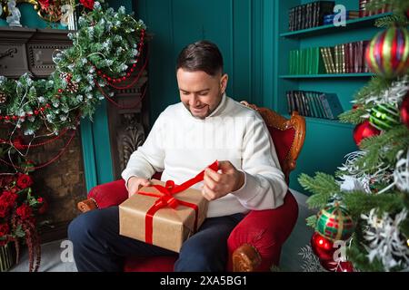 Hübscher junger Mann in weißem Pullover, der zu Hause ein weihnachtsgeschenk eröffnet. Stockfoto