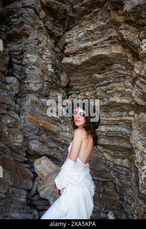 Eine Frau in weißem Kleid und schwarzem Hut steht gegen eine raue Felsformation Stockfoto