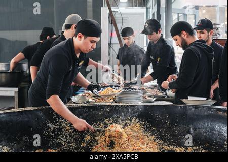 Männliche Köche legen fertige traditionelle usbekische Pilaw aus einem großen Kessel auf Teller im zentralasiatischen Pilaw Center Besh Qozon. Taschkent, Usbekistan - 16. April 2024 Stockfoto