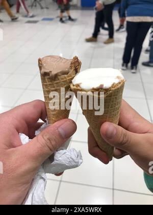 Nahaufnahme einer Hand, die zwei Eiskegel mit schmelzenden Schaufeln hält, drinnen mit verschwommenen Menschen im Hintergrund Stockfoto