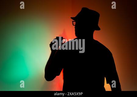 Mann mit Hut in Silhouette hält eine Tasse Kaffee vor bunter Hintergrund. Stockfoto