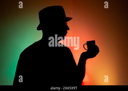 Mann mit Hut in Silhouette hält eine Tasse Kaffee vor bunter Hintergrund. Stockfoto