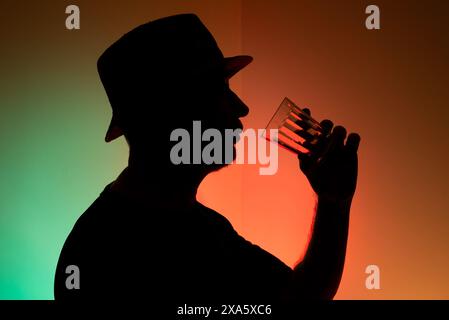 Mann mit Hut in Silhouette, der ein Glas Wasser vor bunter Hintergrund hält. Stockfoto