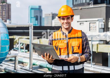 Ein Mann mit Helm, der an einem Laptop in der Nähe von Industrierohren in Thailand, Bangkok, arbeitet Stockfoto