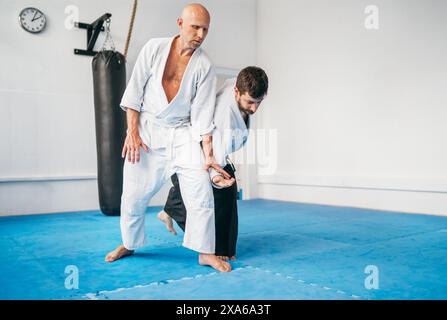 Zwei Männer üben Aikido im Dojo mit traditionellen weißen Kimono und Hakama. Ein Mann führt eine präzise Technik aus, um seinen Partner dazu zu führen, Dämonen zu mattieren Stockfoto