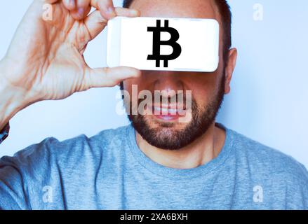 Kaukasischer bärtiger Mann mit dem Handy über den Augen mit Bildschirm mit Bitcoin-Symbol. Stockfoto