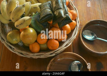 Ein Korbkorb gefüllt mit tropischen Sommerfrüchten wie Passionsfrüchten, Bananen und Orangen auf einem Holztisch neben leeren Holzschalen mit Löffel Stockfoto