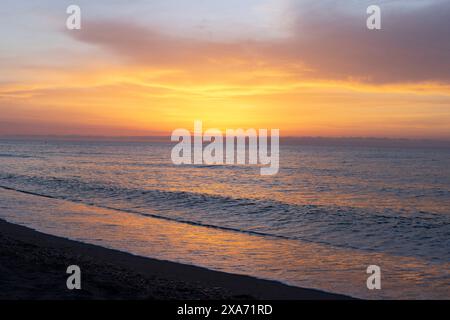 Ein Sonnenaufgang am Strand mit Wolken in leuchtenden Farben, die sich vom Meer reflektieren und eine perfekte Sonnenuntergangsszene für Hintergrundbilder und Urlaubshintergründe schaffen. Stockfoto