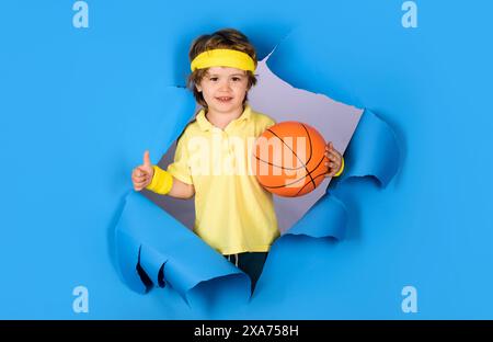 Lächelnder Junge mit Basketballball, der durch das Papierloch schaut und den Daumen nach oben zeigt. Kleiner Basketballspieler in Sportbekleidung mit Ball. Basketballtraining. Stockfoto