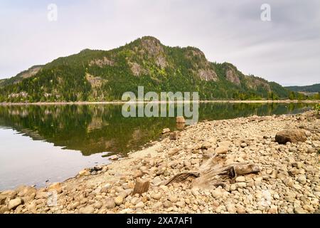 Blick von einem felsigen Kiesstrand auf die steilen Seiten eines gesäumten Berges und seine Reflexion im ruhigen See. Stockfoto