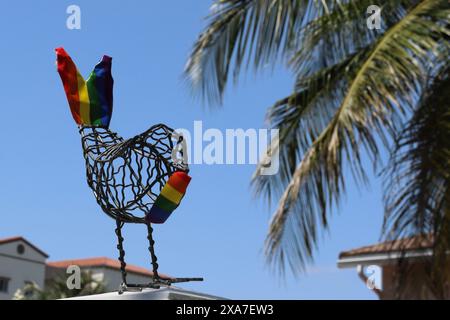 Eine Metallvogelskulptur mit einer regenbogenfarbenen Geschichte und Vorderseite am Strandzaun mit Palmen Stockfoto