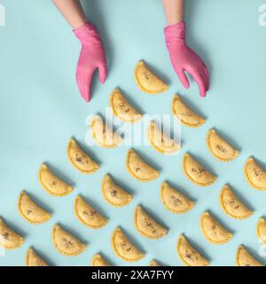 Die Hände eines Bäckers in rosafarbenen Gummihandschuhen legen rohe Samosa auf blauem Hintergrund ab. Traditionelle Bäckerei mit zentralasiatischer Küche, tiefgefrorene Halbfertigprodukte Stockfoto