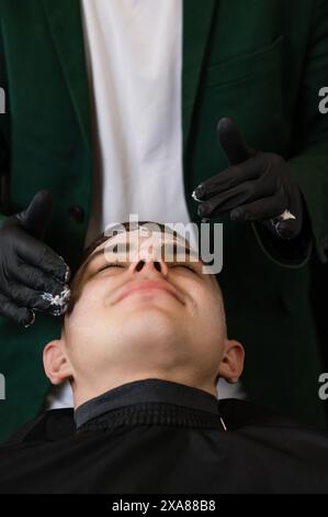 Ein Friseur in schwarzen Handschuhen trägt eine feuchtigkeitsspendende weiße Creme auf das Gesicht des Kunden auf. Eine Kosmetikerin trägt eine weiße Maske mit einem Vitaminkomplex auf die Haut von auf Stockfoto