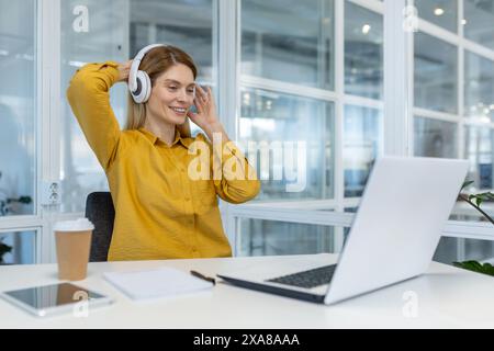 Frau, die Musik über Kopfhörer genießt, während sie an einem Laptop in einer modernen, hellen Büroumgebung arbeitet. Sie wirkt entspannt und glücklich. Stockfoto