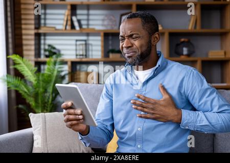 Ein kranker Patient hält seine Brust, während er sich mit einem Arzt über einen Videoanruf aus seinem Wohnzimmer berät. Gesundheitswesen und Telemedizin-Konzept. Stockfoto