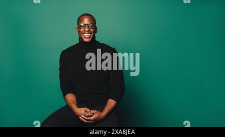 Selbstbewusster afrikanischer Mann mit trendiger Brille und lässigem Pullover. Sitzen im Studio mit modischem grünen Hintergrund. Unbeschwertes Lächeln und eleganter Stil Stockfoto