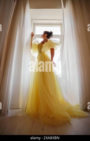 Die Silhouette einer Frau in einem goldenen, luxuriösen Kleid vor dem Hintergrund eines Fensters hält einen Vorhang mit den Händen. Elegante Dame in gelber Seide Stockfoto