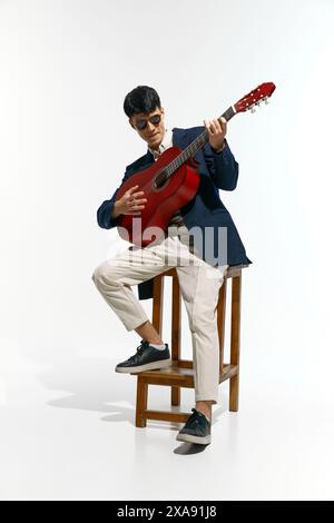 Junger Mann in marineblauem Blazer und beigefarbener Hose, mit Sonnenbrille, sitzt auf Holzhocker und spielt Akustikgitarre vor weißem Studiohintergrund. Stockfoto