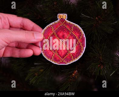 Eine Frauenhand, die ein rotes kreuzgenähtes Weihnachtsschmuck vor einem Weihnachtsbaumhintergrund hält. Stockfoto