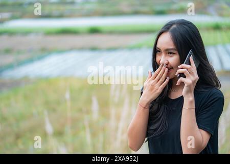 Eine junge Frau mit langen schwarzen Haaren, die ein schwarzes Hemd trägt, lächelt und redet auf einem Smartphone, während sie draußen auf einem Feld steht. Stockfoto