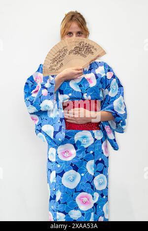 Eine Cosplayer in einem blauen Kimono mit einem roten Flügel, der einen Holzfächer vor ihrem Gesicht hält Stockfoto