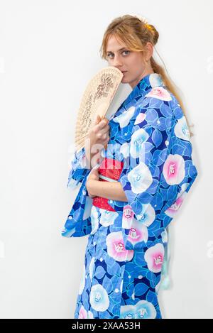 Eine Cosplayer in einem blauen Kimono mit einem roten Flügel, der einen Holzfächer vor ihrem Gesicht hält Stockfoto