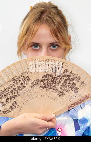 Eine Cosplayer in einem blauen Kimono mit einem roten Flügel, der einen Holzfächer vor ihrem Gesicht hält Stockfoto