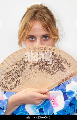 Eine Cosplayer in einem blauen Kimono mit einem roten Flügel, der einen Holzfächer vor ihrem Gesicht hält Stockfoto