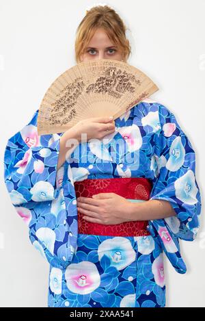 Eine Cosplayer in einem blauen Kimono mit einem roten Flügel, der einen Holzfächer vor ihrem Gesicht hält Stockfoto