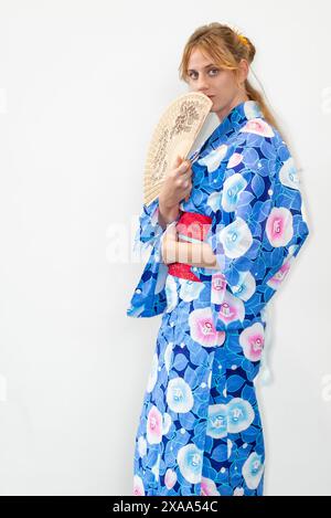 Eine Cosplayer in einem blauen Kimono mit einem roten Flügel, der einen Holzfächer vor ihrem Gesicht hält Stockfoto