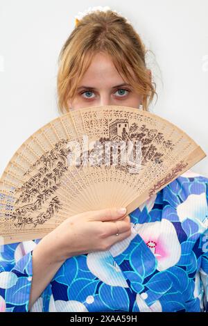 Eine Cosplayer in einem blauen Kimono mit einem roten Flügel, der einen Holzfächer vor ihrem Gesicht hält Stockfoto