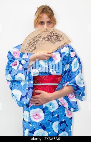 Eine Cosplayer in einem blauen Kimono mit einem roten Flügel, der einen Holzfächer vor ihrem Gesicht hält Stockfoto