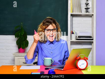 Wütende Studentin, die mit Laptop im Klassenzimmer arbeitet. E-Learning und Bildung. Schreiender Lehrer mit Brille sitzt mit Schoß am Schreibtisch Stockfoto