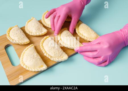 Die Hände eines Bäckers in rosafarbenen Gummihandschuhen legen rohe Samosa auf das Holzbrett. Traditionelle zentralasiatische Küche, gefrorene Halbfertigprodukte Stockfoto