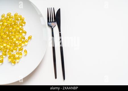 Viele klare gelbe Fischöl-Pillen, Omega 3 auf einem weißen Teller mit Gabel und Messer auf hellem Hintergrund. Nahaufnahme. Kopierbereich. Hochwertige Fotos Stockfoto