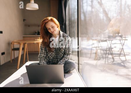 Porträt einer glücklichen freiberuflich arbeitenden Frau, die an einem Laptop arbeitet und auf einer breiten Fensterbank mit Blick auf die Natur an sonnigem Tag sitzt. Stockfoto