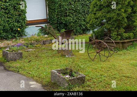 Ein altes rostiges Anbaugerät sitzt auf dem Gras vor einem Haus mit grünem Sträucher. Stockfoto