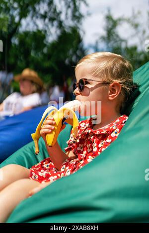 Ein kleines Mädchen isst eine Banane. Porträt eines modischen, stilvollen, schönen Mädchens in Sonnenbrille und rotem Kleid, während er in einem Sitzsack-Stuhl sitzt Stockfoto