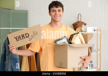 Junger Mann, der Pappe hält, mit BRIEFMARKENVERKAUF und Kiste im Zimmer mit unerwünschtem Zeug Stockfoto