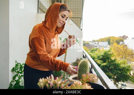 Eine lateinische glückliche Frau in einem orangefarbenen Hoodie pflegt ihre Pflanze auf einem Balkon, während sie Mate trinkt. Die Pflanze ist ein Kaktus und die Frau lächelt und qu Stockfoto