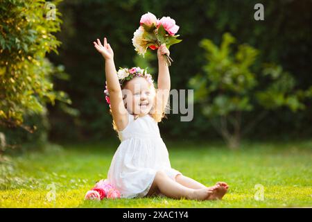 Kleines Mädchen mit Blumenstrauß im sonnigen Garten. Blühender Sommerpark. Kind mit Blumengeschenk für Geburtstagsfeier oder Osterfeier. Stockfoto