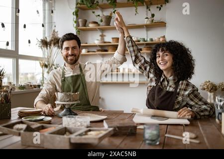 Lächelndes Paar im Töpferstudio mit Schürzen und High-Fiving, während er mit Töpferwerkzeugen an einem Arbeitstisch sitzt. Stockfoto
