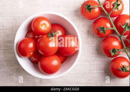 Rote Kirschtomaten in einer weißen Schüssel auf Leinenstoff. Frische, reife Art kleiner und runder Cocktailtomaten, links noch an der Rebe. Stockfoto