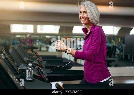 Seniorin, auf Laufband und Lächeln für Musik im Fitnessclub, Wasser und Flasche für Gesundheit im Wellness. Reife, weibliche Läuferin und glücklich mit Stockfoto