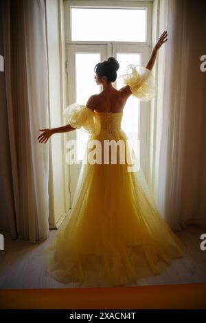 Die Silhouette einer Frau in einem goldenen, luxuriösen Kleid vor dem Hintergrund eines Fensters hält einen Vorhang mit den Händen. Elegante Dame in gelber Seide Stockfoto