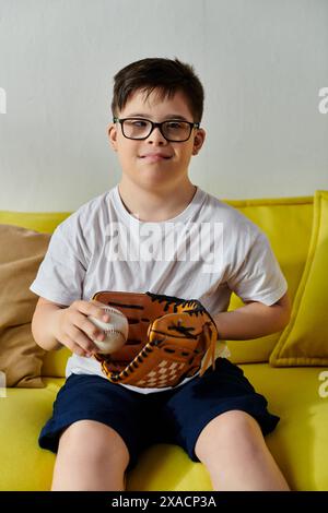 Entzückendes Kind mit Down-Syndrom mit Brille, die auf einer gelben Couch sitzt und einen Baseball hält. Stockfoto