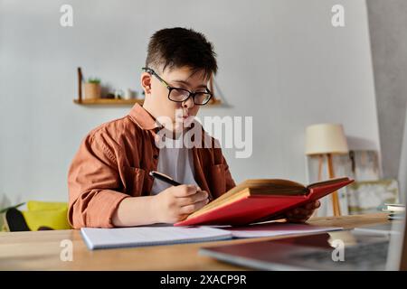 Ein Junge mit Down-Syndrom liest ein Buch und arbeitet an einem Laptop am Schreibtisch. Stockfoto